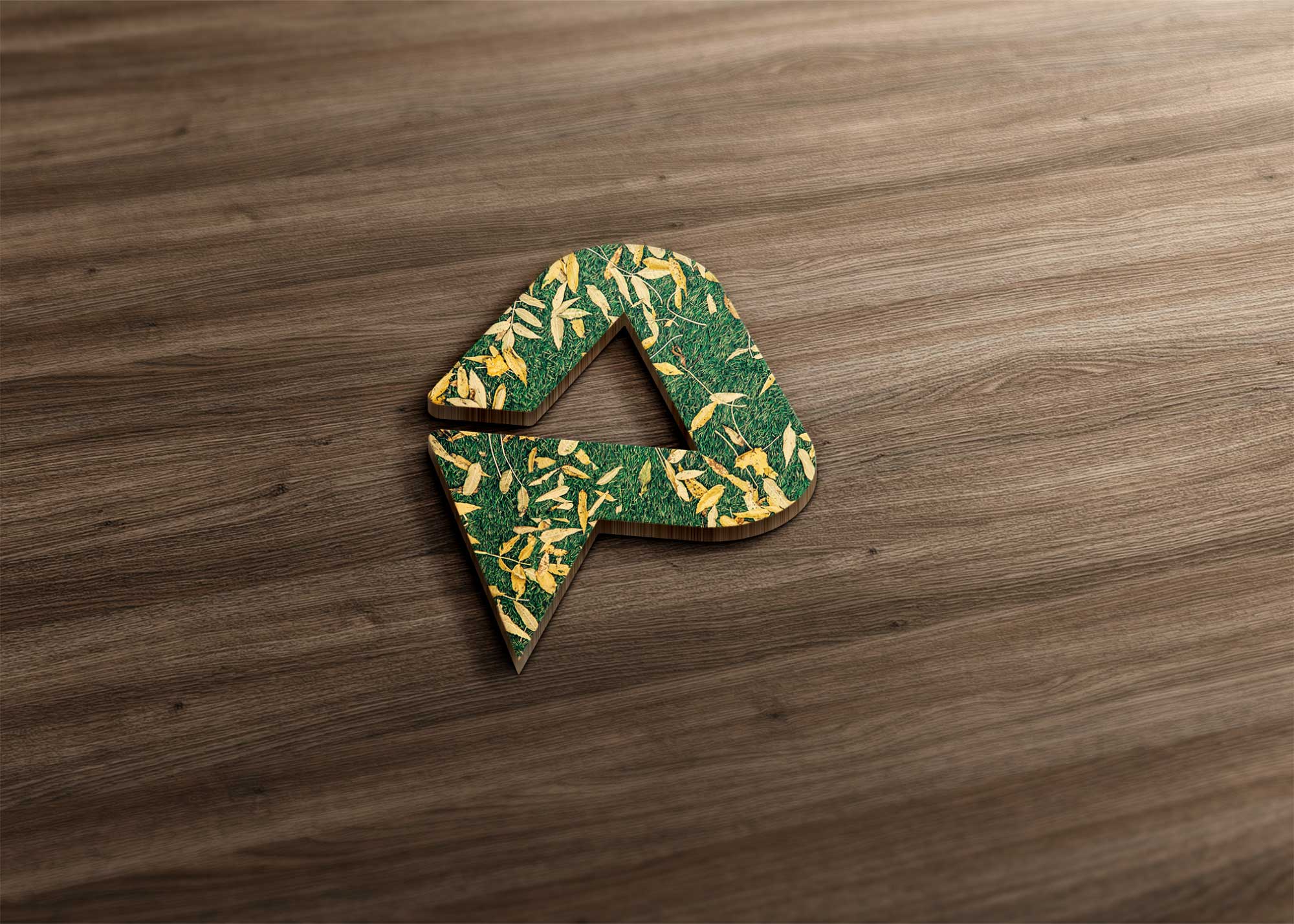 A 3D logo resembling the letter "A," covered in green grass and yellow leaves, rests on a wooden surface.