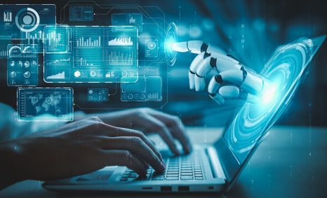 A robotic hand reaches toward a glowing digital interface while a person types on a laptop, surrounded by data visualiza