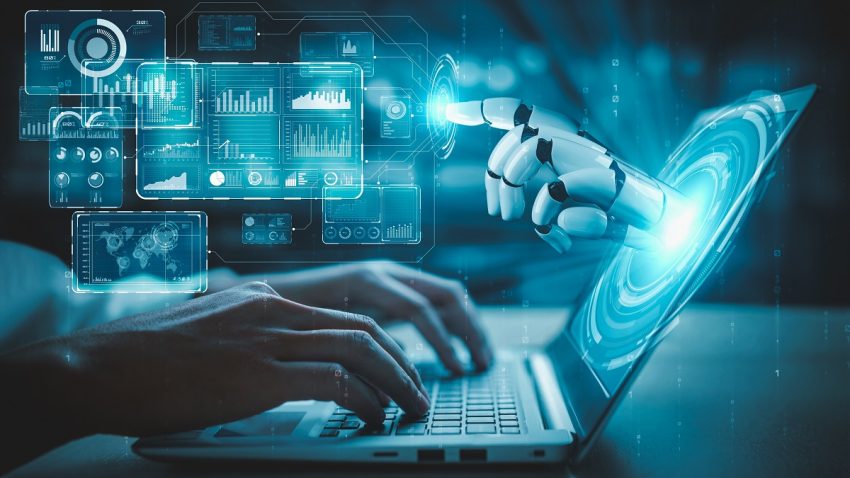A robotic hand reaches toward a glowing digital interface while a person types on a laptop, surrounded by data visualiza