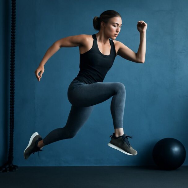 A woman in a black tank top and leggings leaps energetically against a blue wall, showcasing her athleticism.